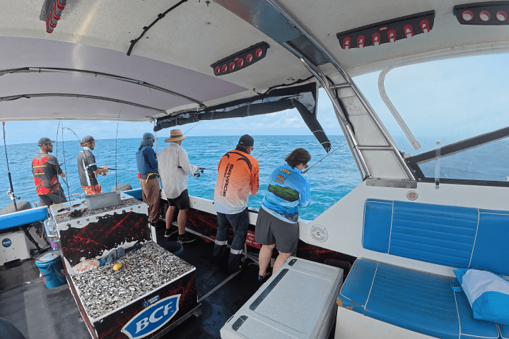 Five people fishing on a boat wearing colorful shirts, with ocean in the background.
