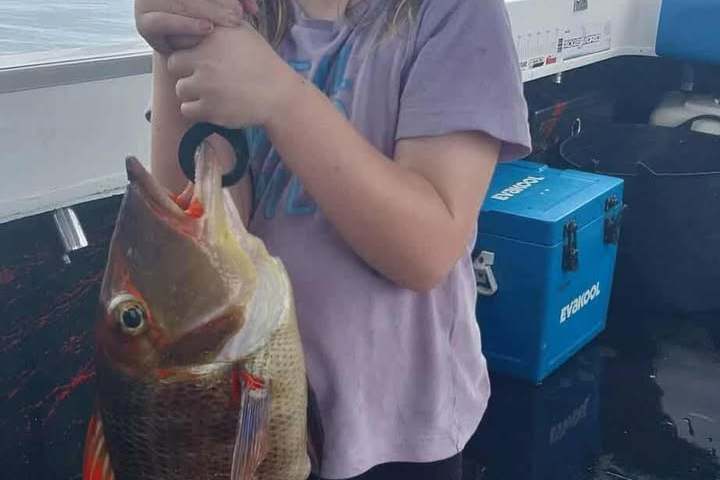 Child on a boat holding a large fish with a fishing rod, smiling, with cloudy sky in the background.