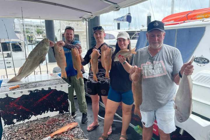 Four people on a boat dock holding freshly caught fish, with boats in the background.