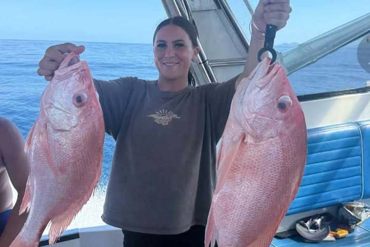 Person on a boat holding two large fish, one in each hand, against a sunny ocean backdrop.