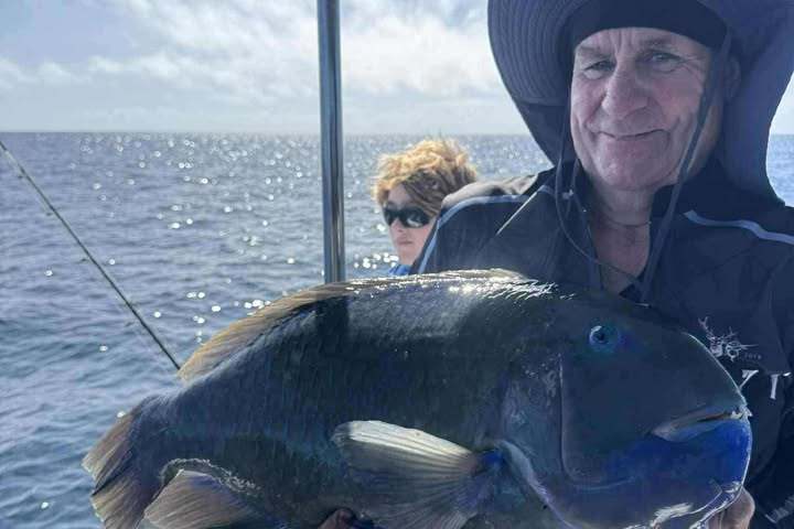 Man in hat holding a large fish on a boat, with ocean in the background.