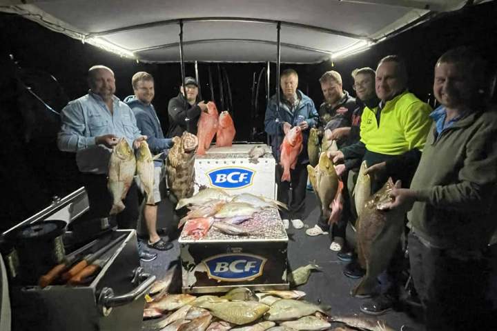 Group of people on a boat holding various fish at night with more fish on the deck.