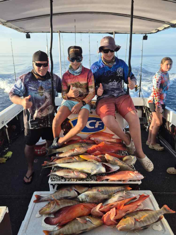 Three people on a boat with a large catch of colorful fish, giving thumbs up.