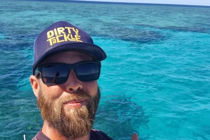 Person in sunglasses, cap, and beard, taking a selfie with clear ocean water in the background.
