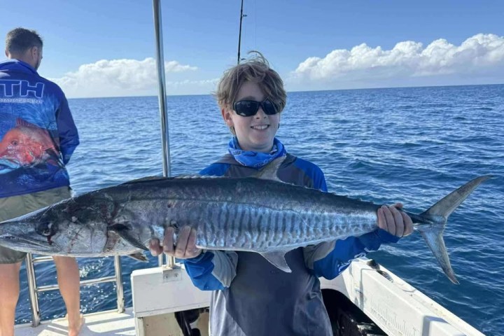 Person holding a large fish on a boat, with another person facing the ocean in the background.